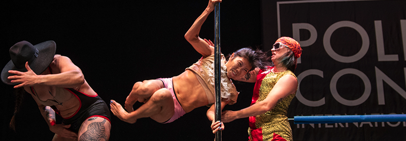 A Pole Dancer Swings Horizontally Around A Pole And A Performer Ducks As If About To Get Kicked.