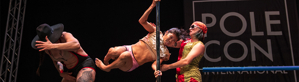 A Pole Dancer swings horizontally around a pole and a performer ducks as if about to get kicked.